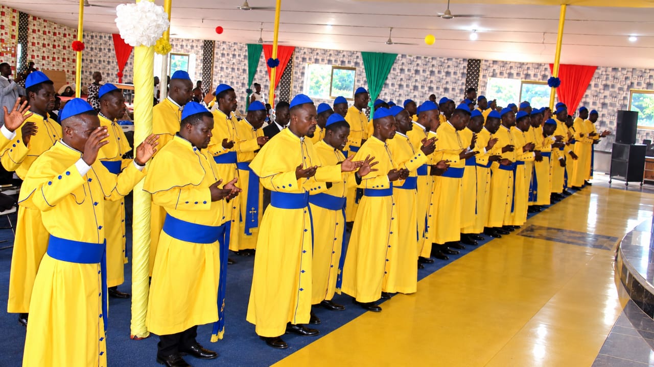 Rows of graduands standing in yellow and blue robes with hands raised in prayer during the service