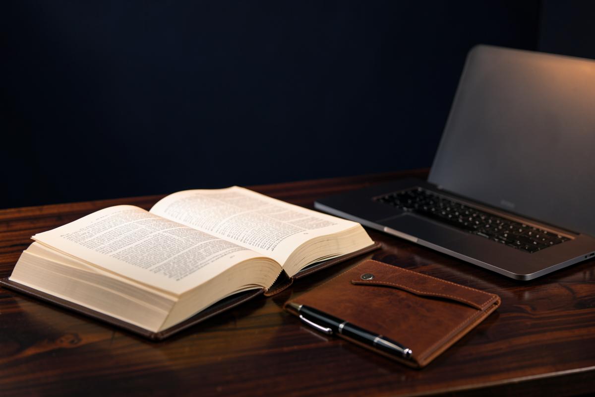 An open Bible, a leather notebook and a laptop on a dark wooden desk, representing study and church administration